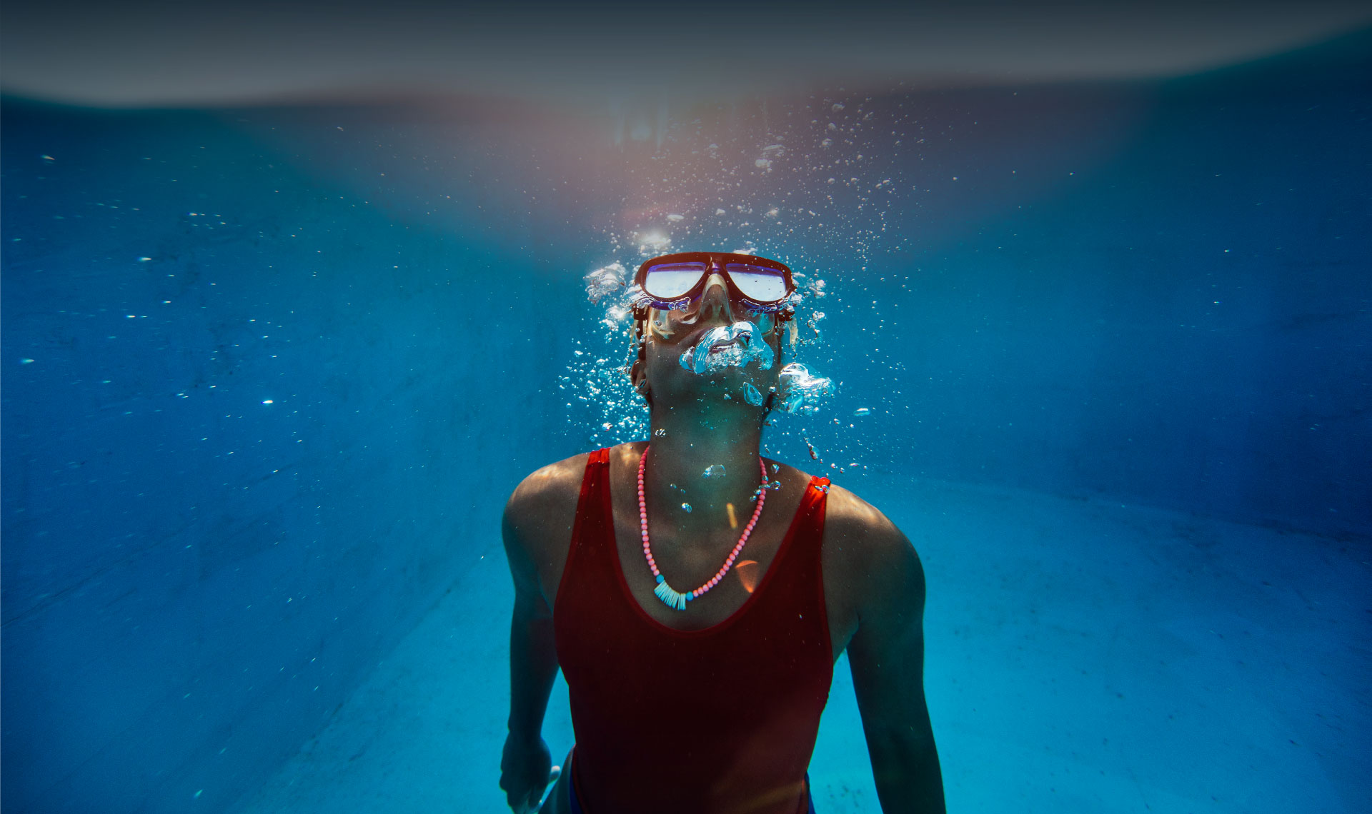 Woman wearing a scuba mask swimming to the surface of a pool while blowing bubbles. 