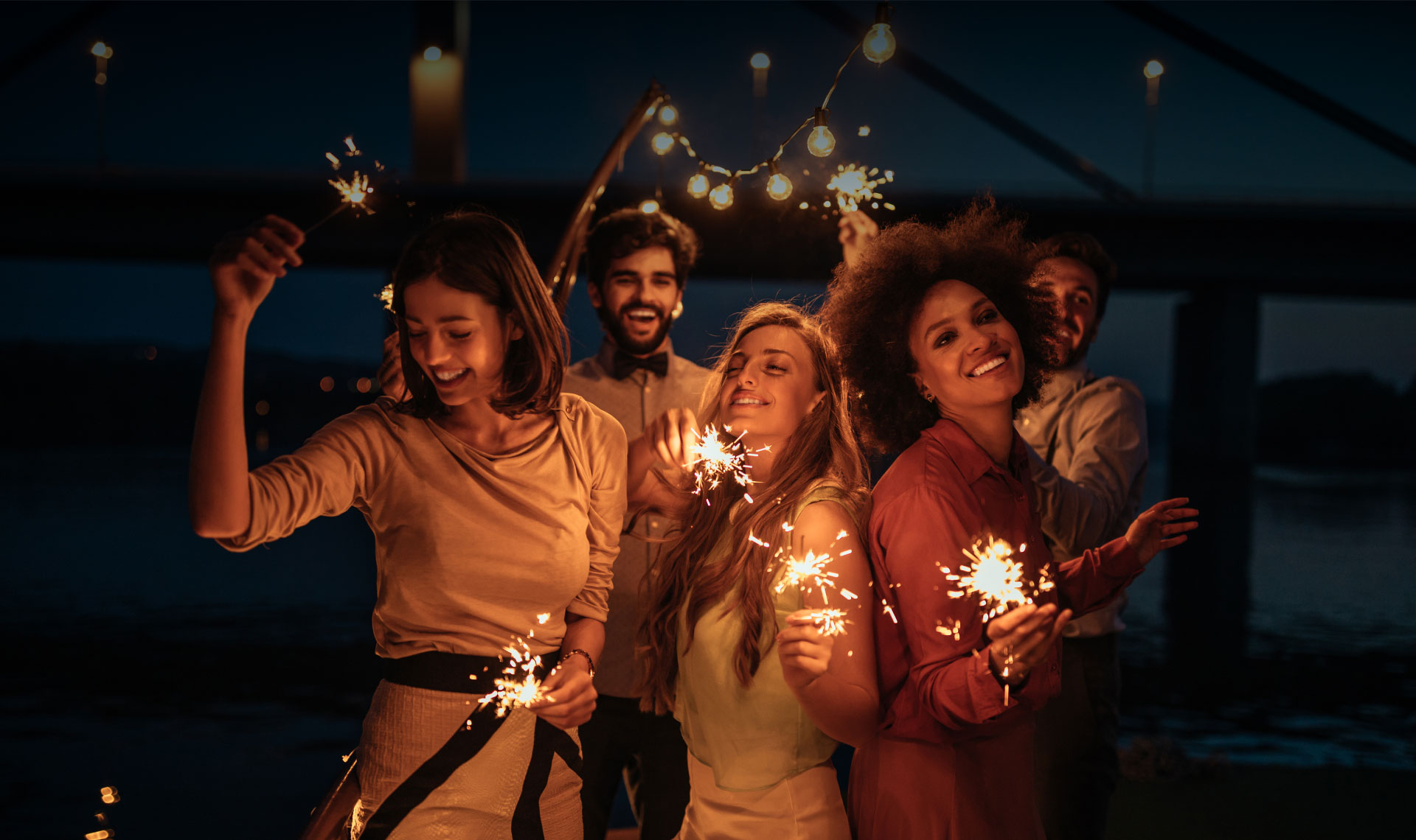 Five friends with sparklers dancing on the beach at night.